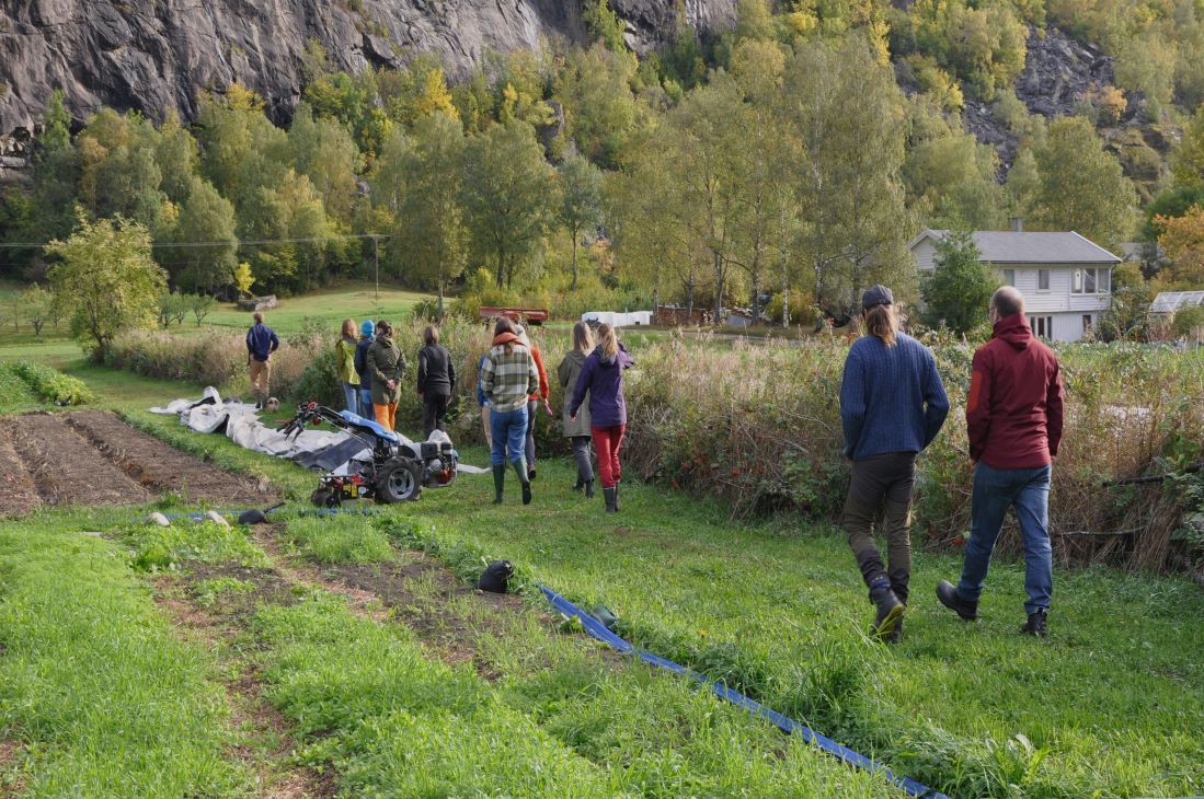 Personer som går langs en åkerkant med hus og fjell i bakgrunnen.