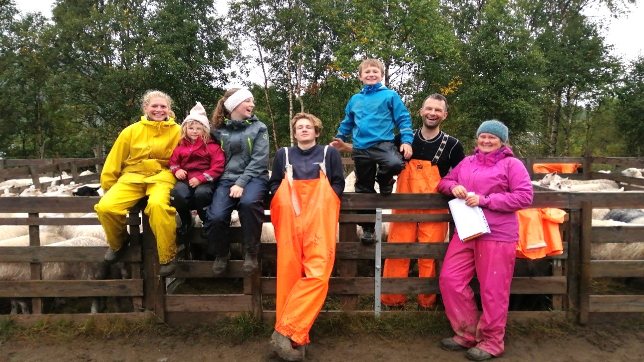 7 personer lener seg mot et gjerde, de har på seg regntøy. Trær i bakgrunnen