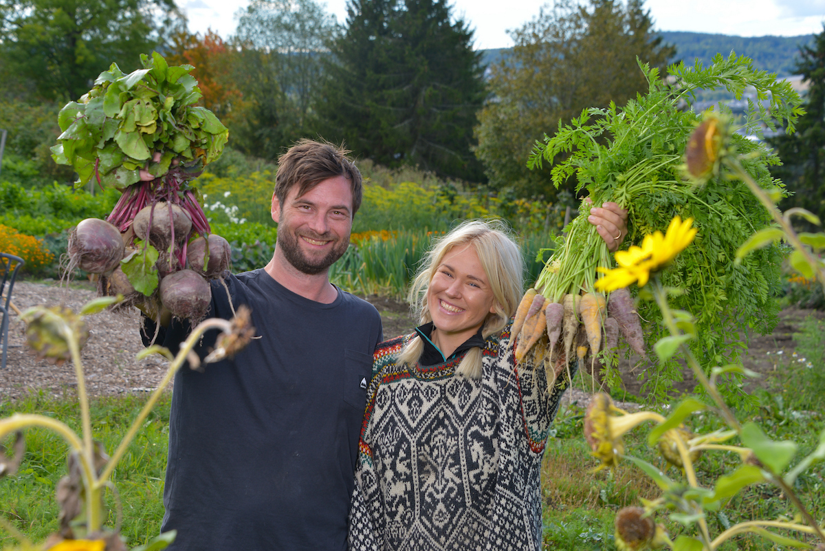 To personer som smiler og holder grønnsaker i bunter opp i hendene