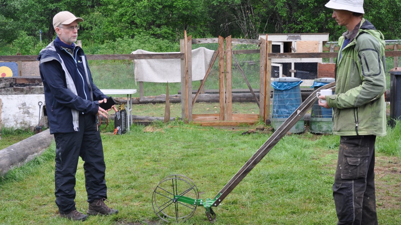 To menn som står og holder i et hageredskap og snakker med hverandre. Hageutstyr i bakgrunnen.