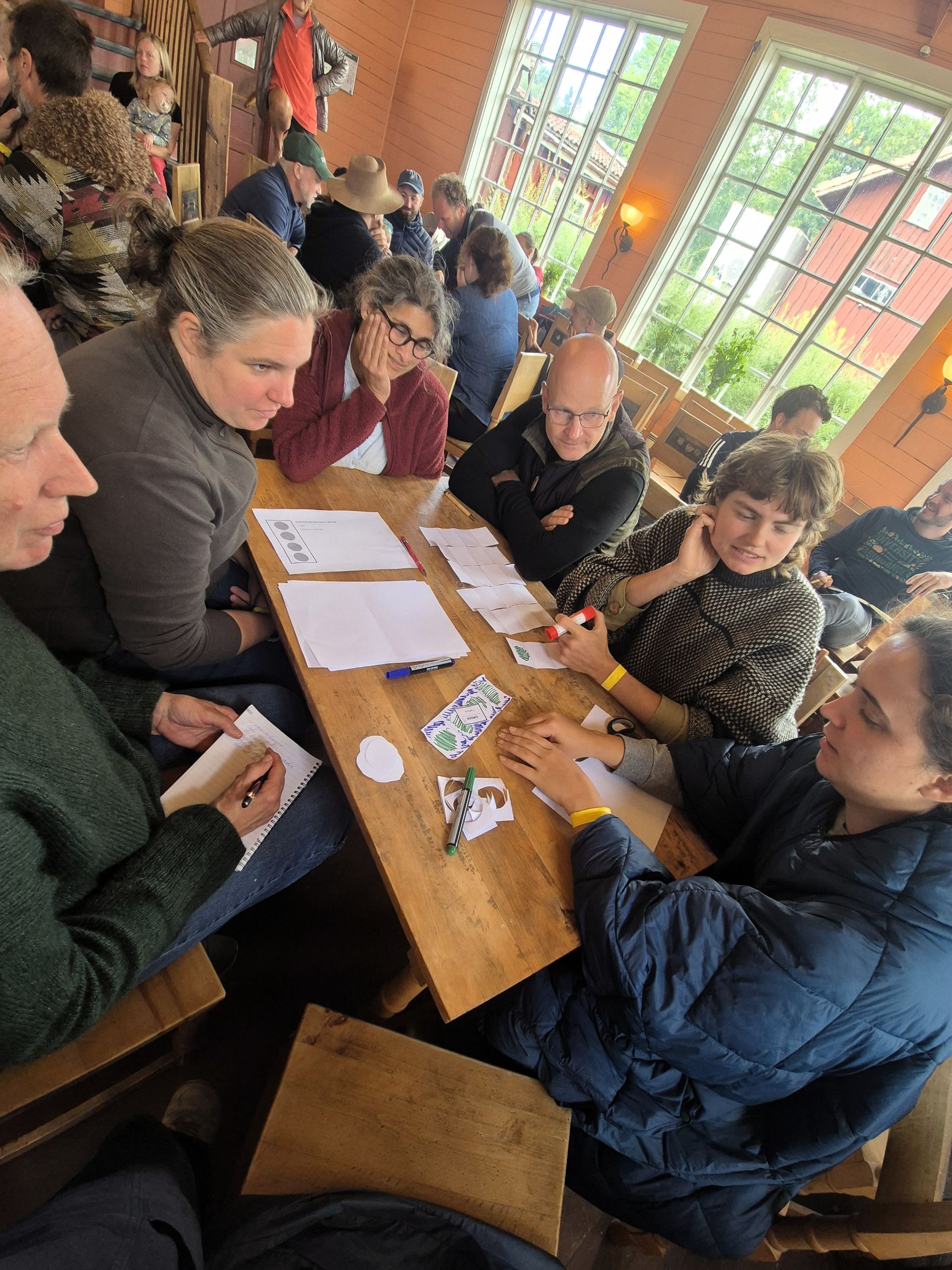 Personer rundt et bord og i bakgrunnen, som diskuterer noe ved et bord