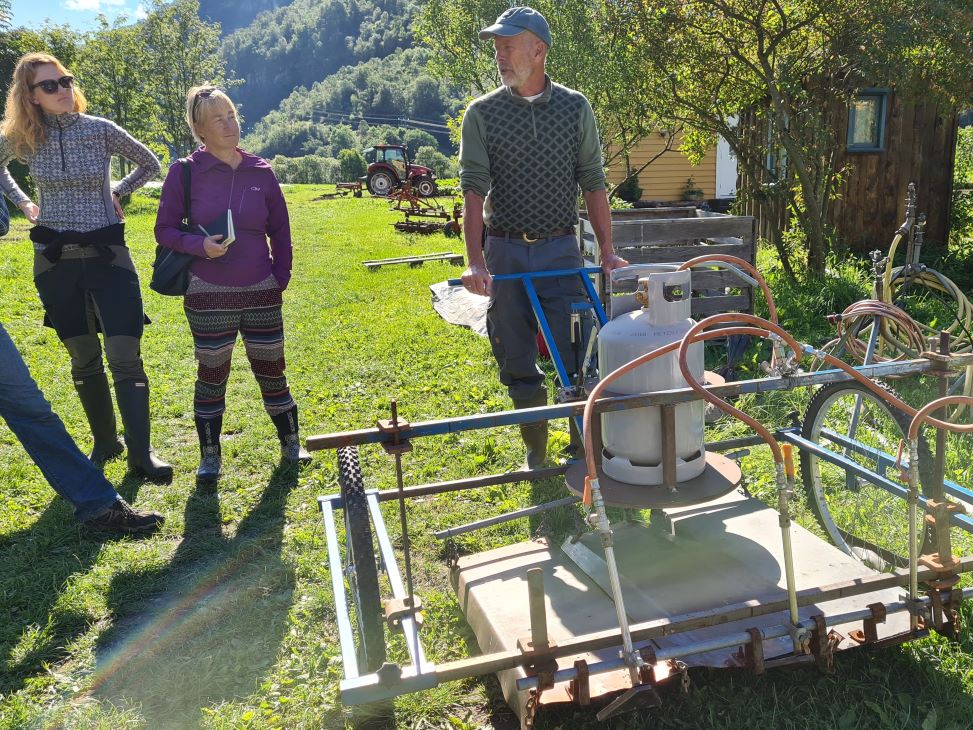 Mann med to damer til venstre som ser på en ugrasbrenner, stativ på hjul med gassdunk. I natur.