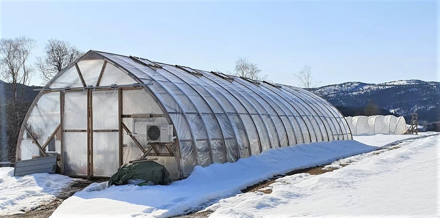 Tunell med stålstenger dekt med lys plast og svart plast med steiner på bakken. Et hus i Bakgrunnen.