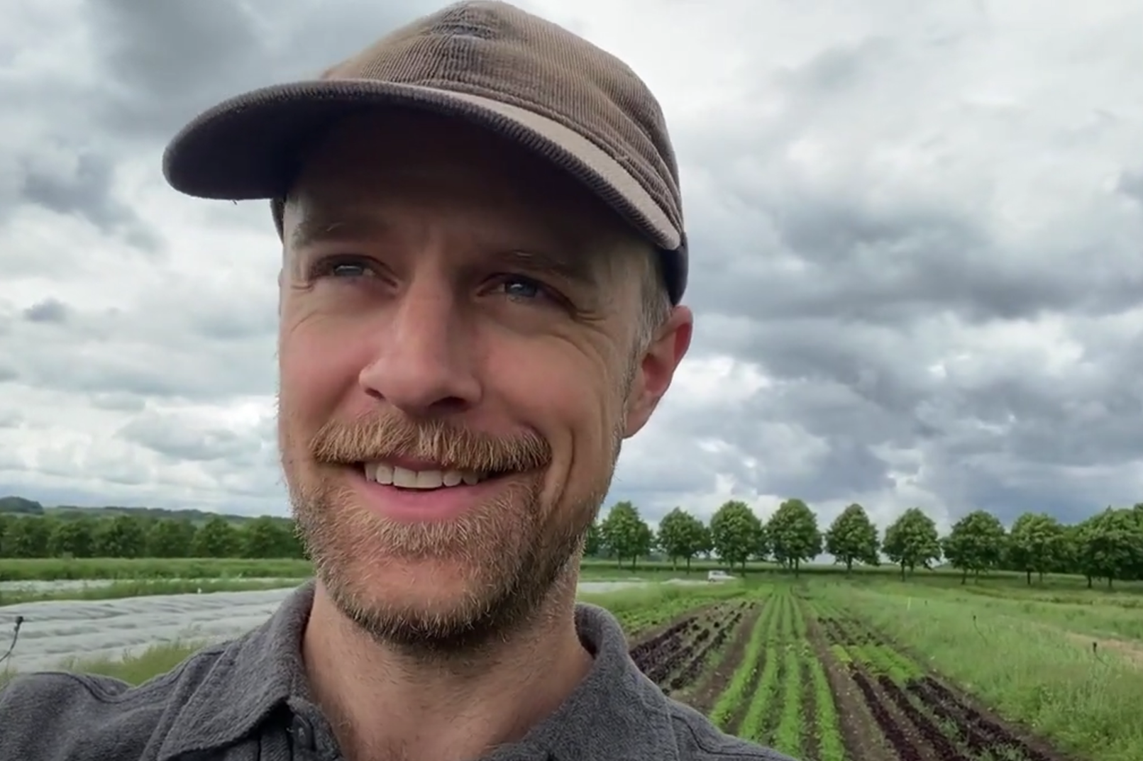 mann med caps på en åker med grønnsaker (rader) og trær i bakgrunnen. 