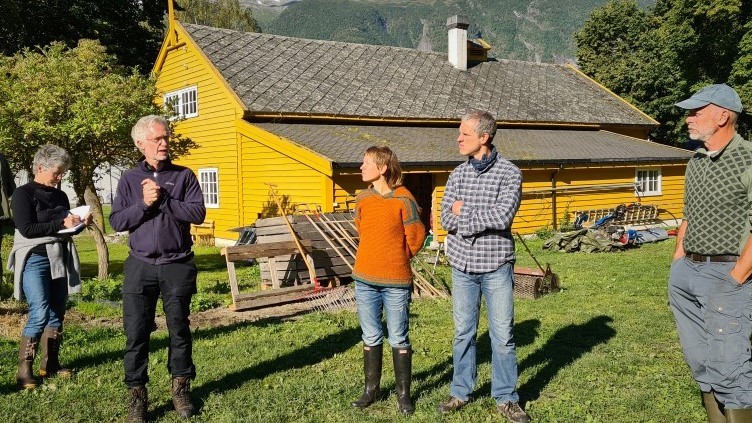 Ulike bønder på markvandring på jordet, ved et gult hus. 