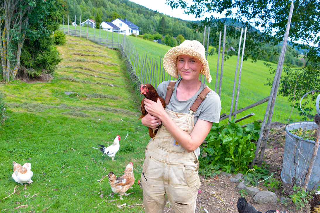 Kvinne med hatt og en høne i armkroken, ute på et jorde. 