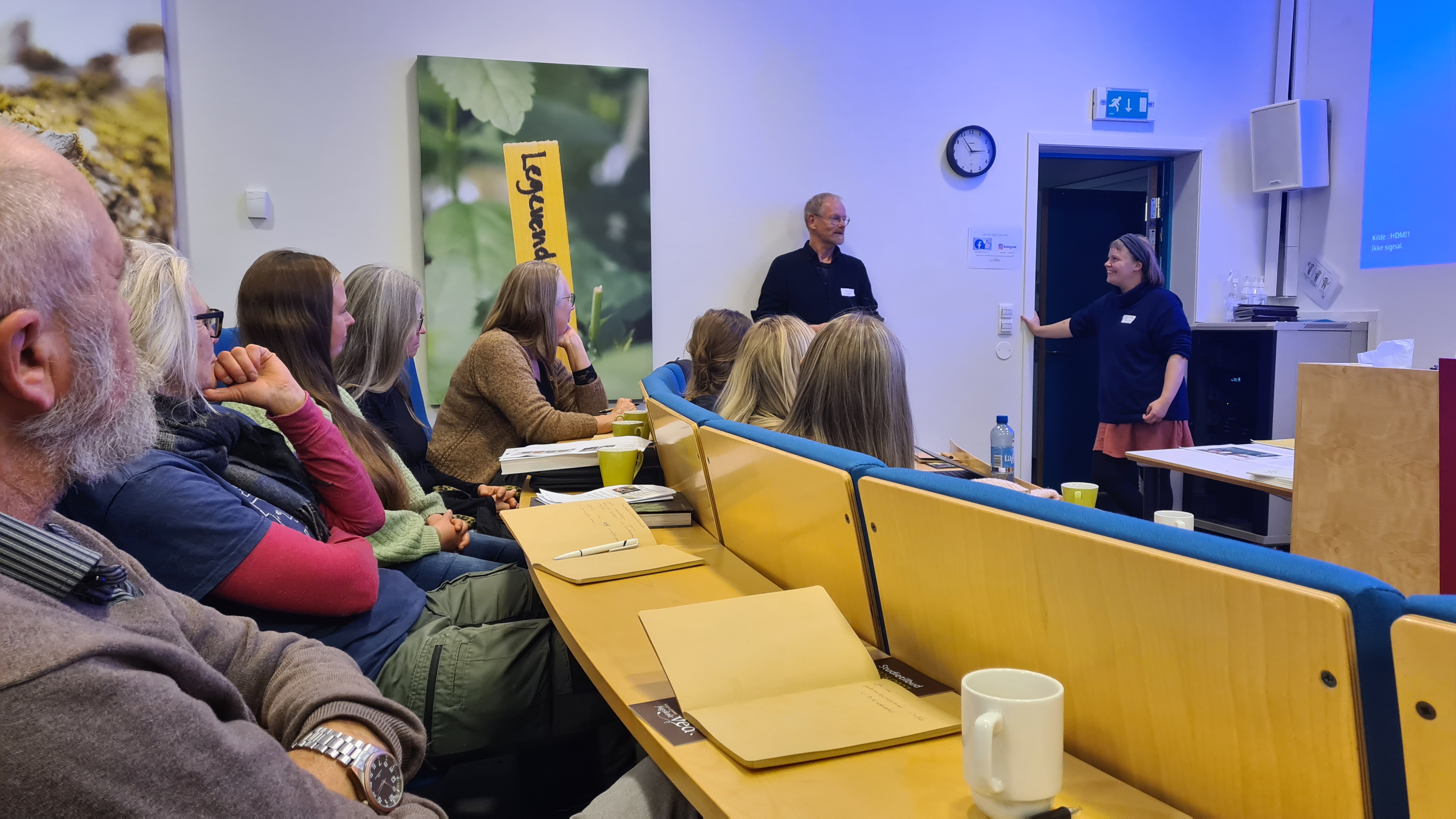 Bildet er tatt i et auditorium hvor en mann og en dame prater engasjert for forsamlingen. Vi ser bakhodene til de på første og andre rad.